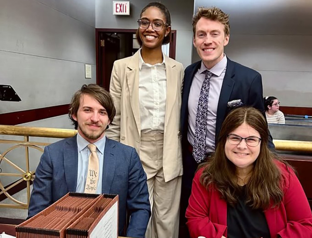 Four students dress in business attire sitting and standing while smiling.